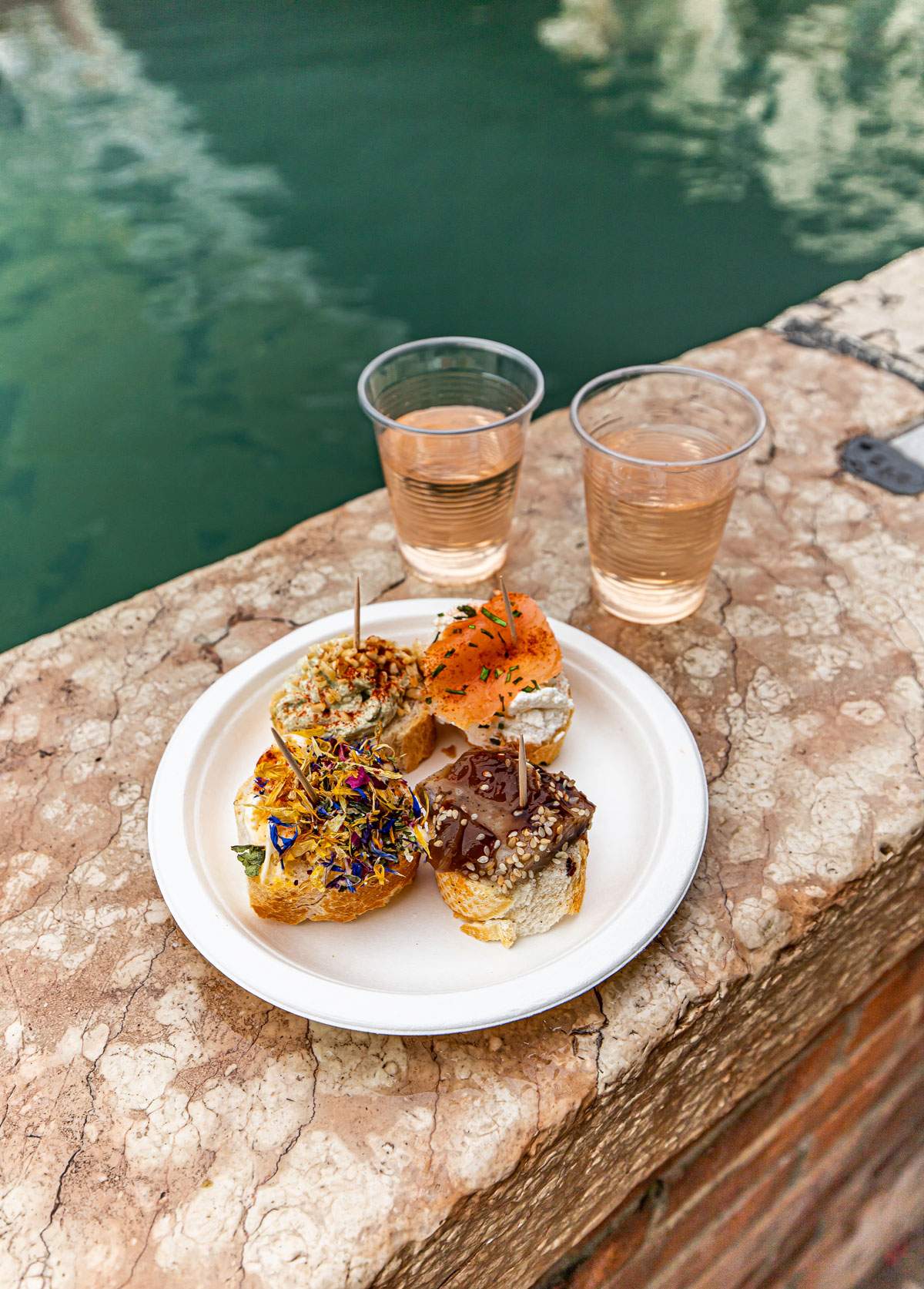 plate of Cicchetti and two cups with wine sat on a ledge by a canal in venice