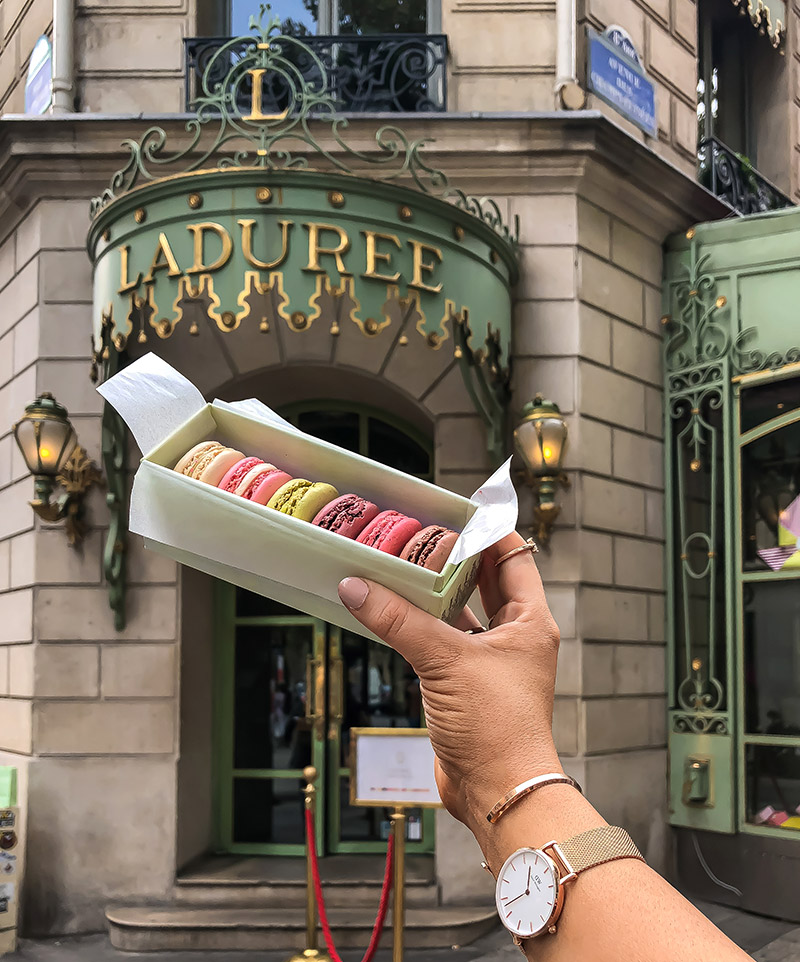 hand holding box of colourful macaroons in front of Laduree cafe