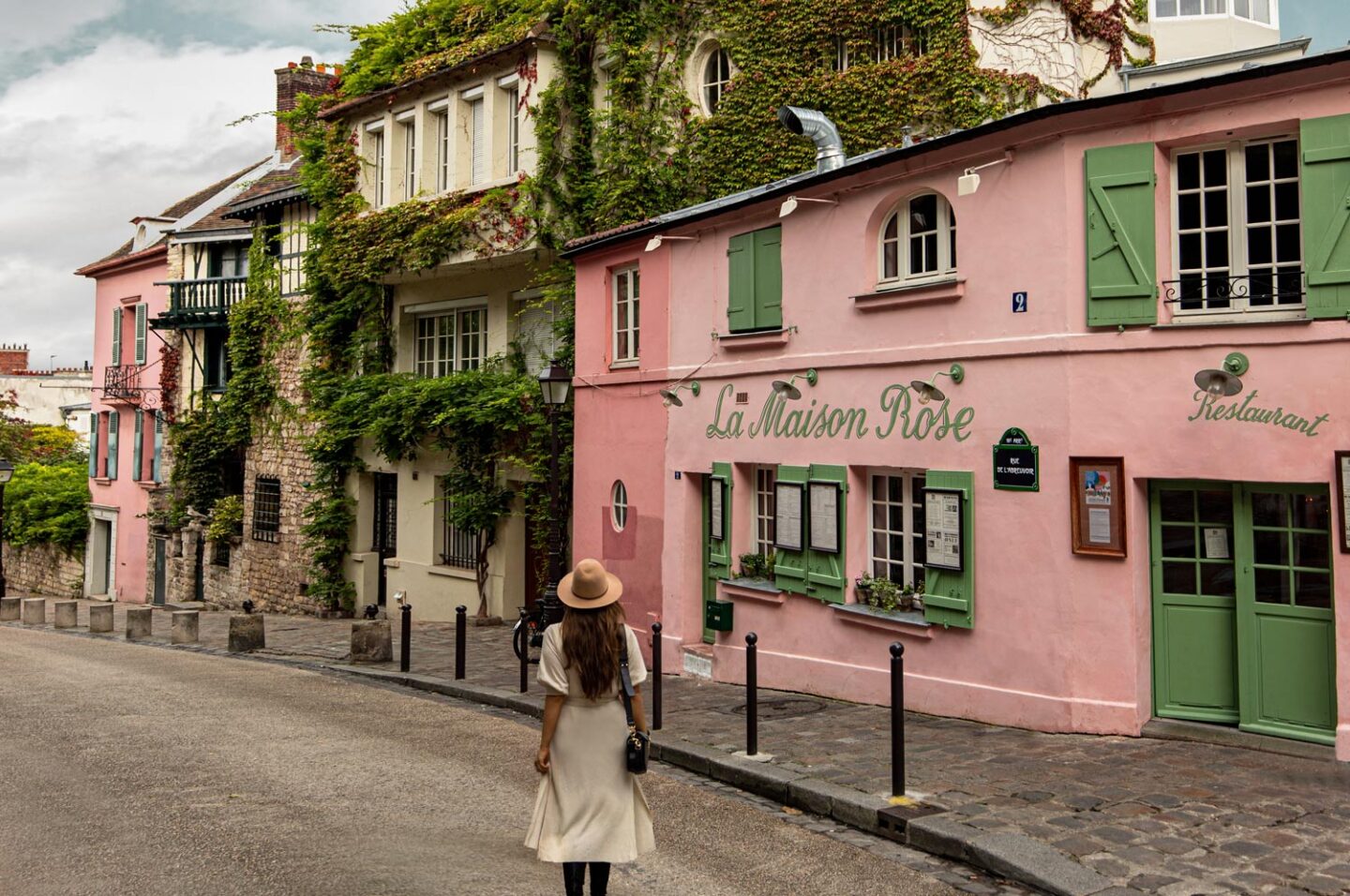 A woman wearing a beige dress and hat walks down a charming, cobblestone street in Paris, approaching the iconic La Maison Rose restaurant. The building features a distinctive pink facade with green shutters, adorned with ivy and plants. The street is lined with other picturesque buildings, creating a quaint and picturesque scene perfect for capturing the essence of Parisian charm.