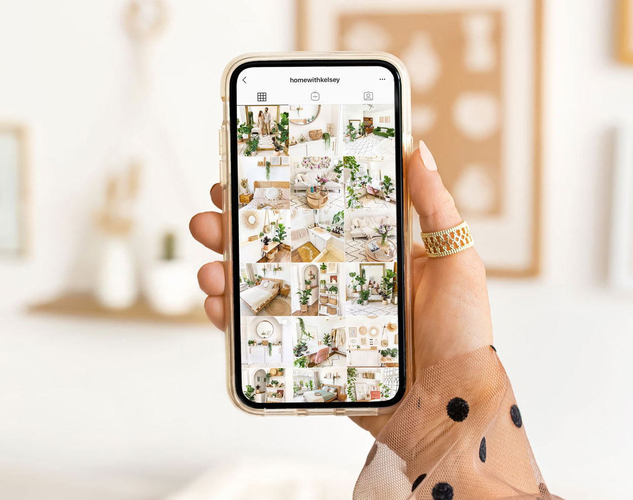 A close-up of a hand holding a smartphone displaying an Instagram profile page. The profile features a grid of aesthetically pleasing interior design photos, showcasing stylish, cozy, and well-decorated rooms with neutral tones and greenery. The person holding the phone is wearing a gold ring and has a light peach nail polish. The background is softly blurred, featuring a bright, minimalist room with subtle decor elements that complement the theme of the Instagram feed. The overall image conveys a sense of modern elegance and tasteful design.






