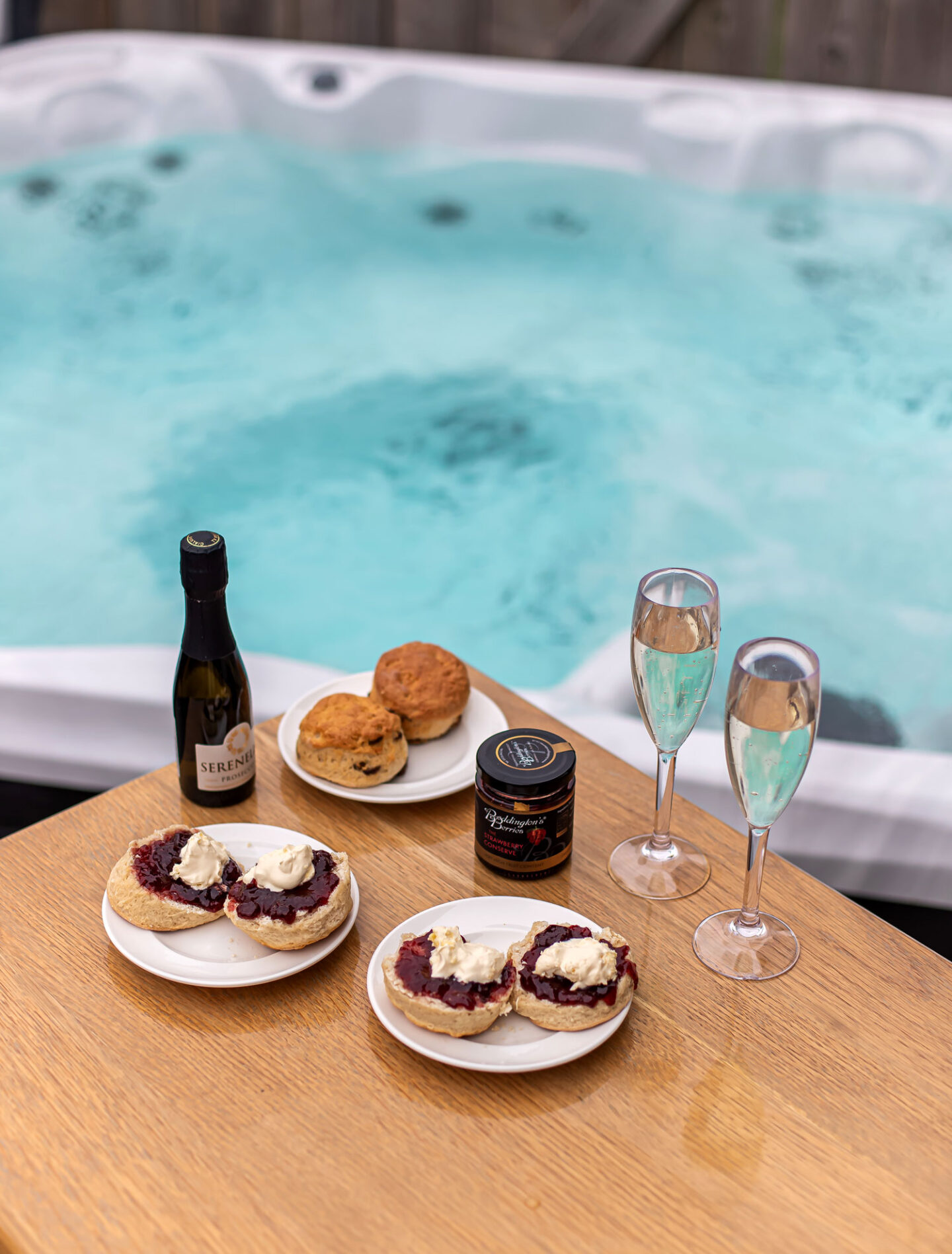 A delightful setup of afternoon tea is placed on a wooden table beside a bubbling hot tub. The table features plates of scones topped with clotted cream and jam, a jar of strawberry jam, a bottle of Prosecco, and two filled champagne flutes. In the background, the inviting turquoise water of the hot tub creates a serene and relaxing atmosphere.