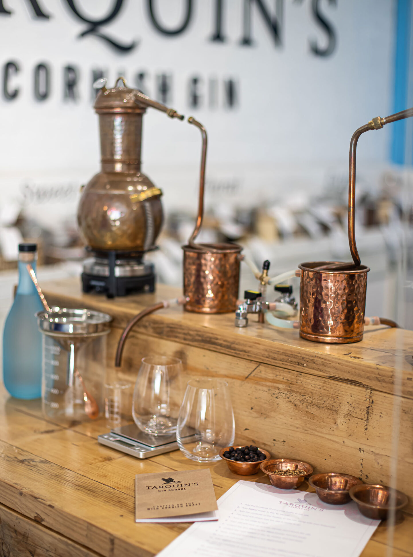 A detailed setup at Tarquin's Cornish Gin distillery showcasing copper stills used for gin making. On the wooden table are various distilling equipment including glass beakers, gin glasses, and small copper bowls filled with botanicals. A booklet titled "Tarquin's Gin School" and a recipe sheet are also visible, indicating an educational and interactive gin-making experience. The background features the distillery's logo, emphasizing the artisanal nature of the process.