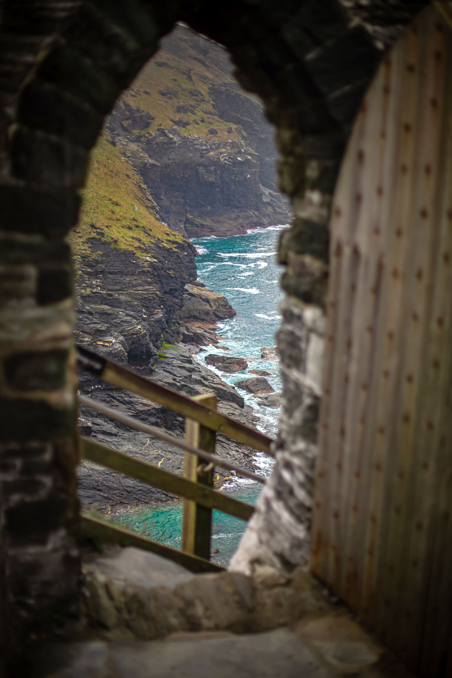 A picturesque view of the rugged Cornwall coastline, seen through the archway of an old stone structure. The turquoise waters crash against the rocky shore, framed perfectly by the weathered walls and wooden door. This perspective offers a sense of history and natural beauty, capturing the essence of Cornwall's dramatic seascape.