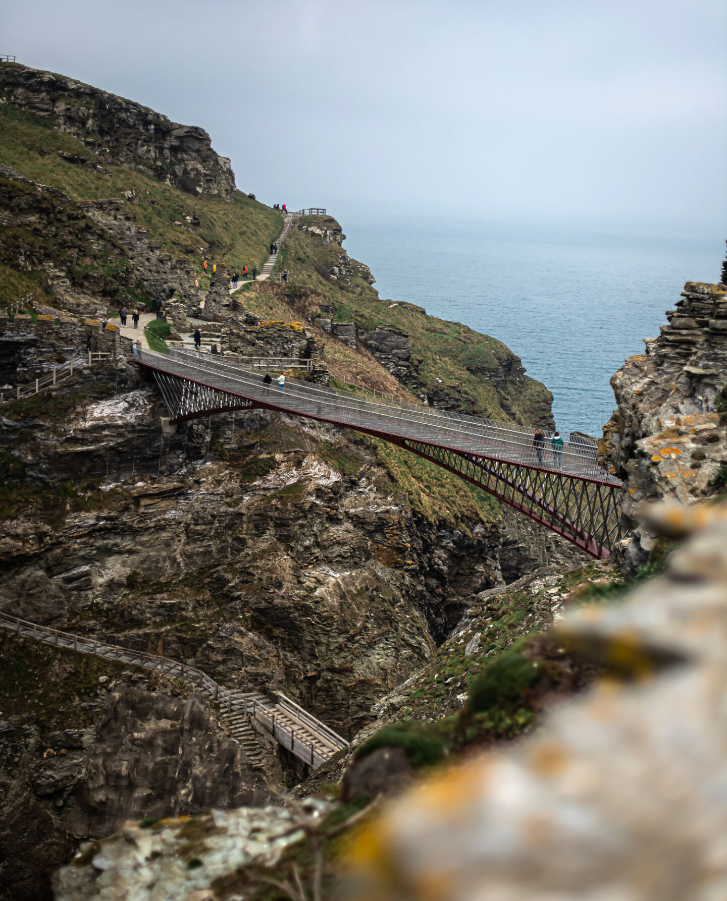 A scenic view of the Tintagel Bridge in Cornwall, stretching across a deep rocky chasm with the sea visible in the background. The modern footbridge, made of metal and wood, connects two cliff tops, allowing visitors to walk between them. People are seen walking along the bridge and the surrounding pathways, highlighting the dramatic and breathtaking landscape of the Cornish coastline.