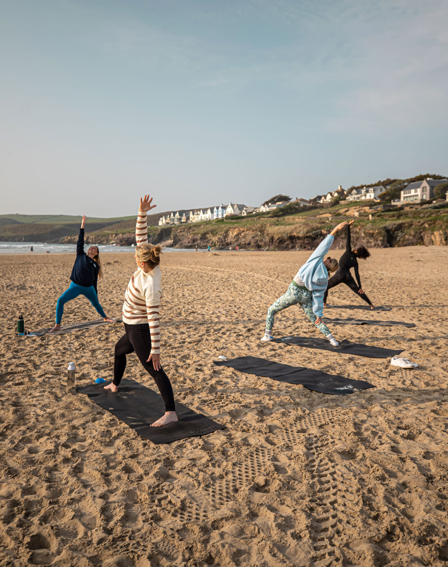 A group of people practicing yoga on a sandy beach in Cornwall, performing stretches and poses on yoga mats. The beach is expansive and tranquil, with gentle waves in the background and picturesque cliffs topped with charming houses. The clear sky and calm atmosphere create an ideal setting for a relaxing and invigorating outdoor yoga session.