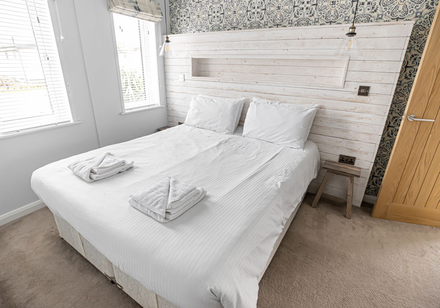 A bright and cozy bedroom featuring a neatly made double bed with crisp white linens and two sets of folded towels. The room has a rustic yet modern design with a whitewashed wooden headboard and decorative wallpaper. Natural light streams in through two large windows, creating a serene and inviting atmosphere.