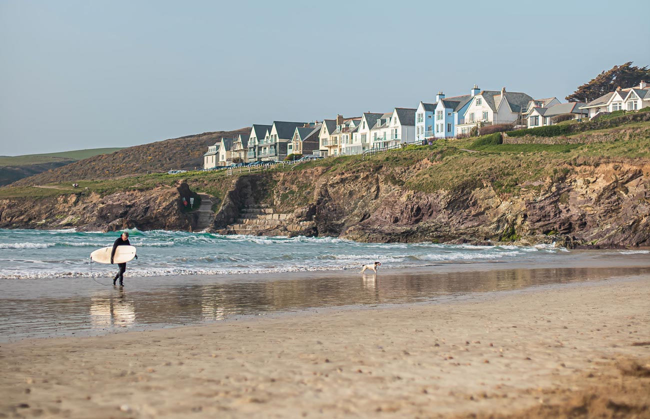 A beautiful beach in Cornwall featuring golden sands and clear blue waters. The coastline is dotted with rugged cliffs and green vegetation, with a few people enjoying the serene, sunny day. The tranquil scene captures the natural beauty and peaceful atmosphere of Cornwall's beaches.