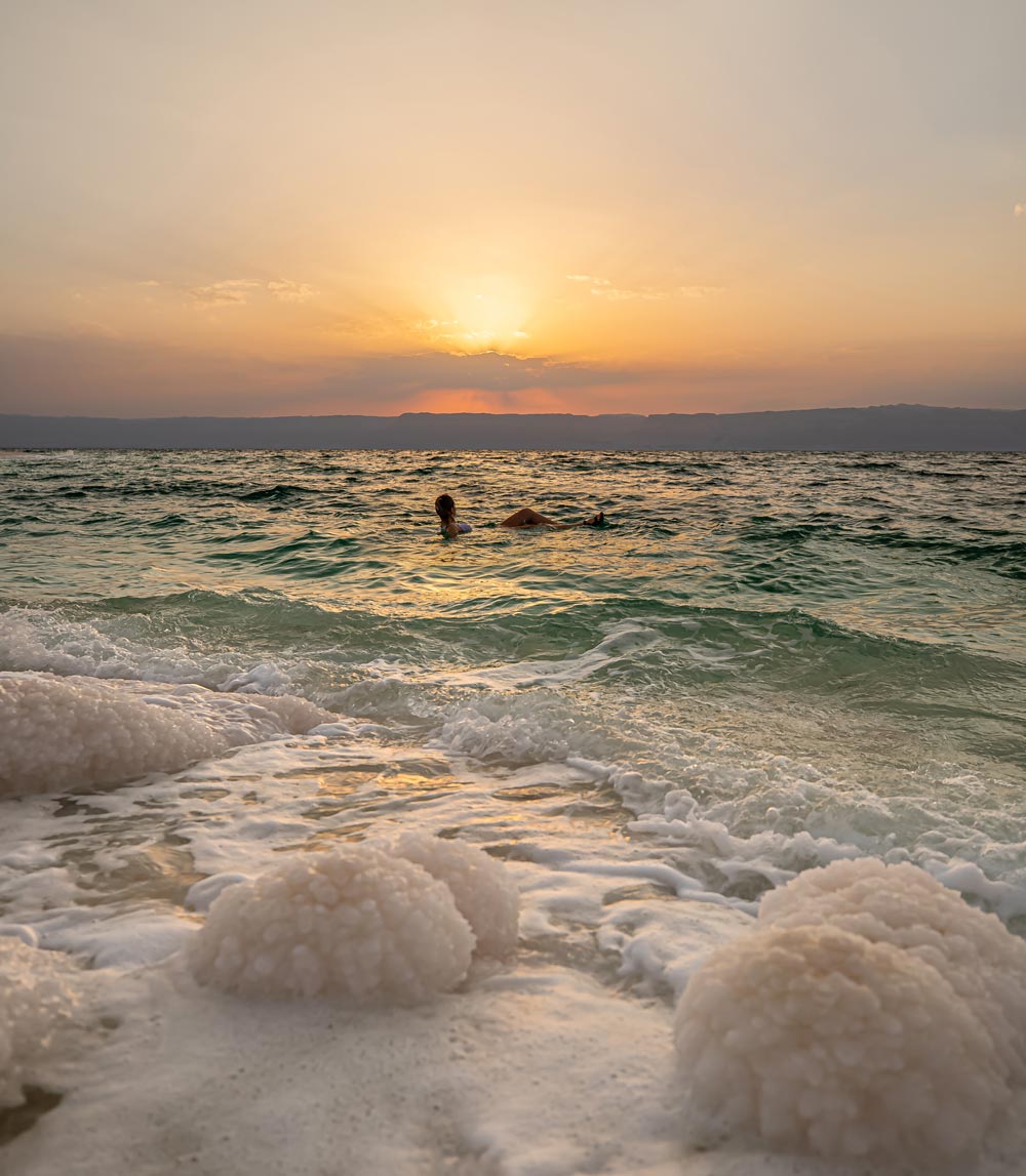 woman floating effortlessly in the dead sea water, the shoreline consists of salt rock formations, the sky is orange as it sets