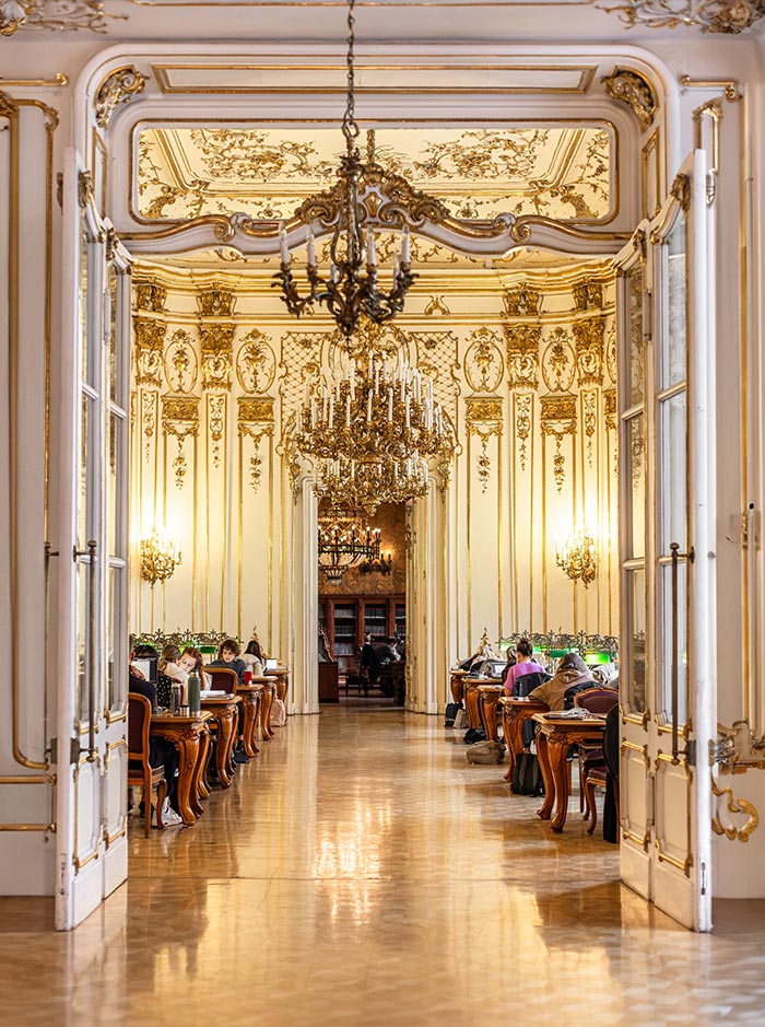 The opulent interior of the Ervin Szabó Library in Budapest, showcasing an ornately decorated reading room with high ceilings, intricate gold accents, and large chandeliers. Rows of wooden tables with green reading lamps are occupied by patrons engrossed in their work. The elegant design and lavish details create a refined, historic atmosphere.