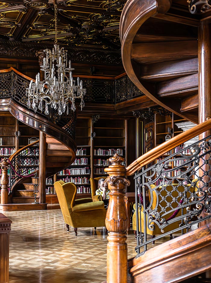 The interior of the Ervin Szabó Library in Budapest, featuring an elegant spiral staircase with intricate wrought-iron railings and dark wood finishes. A large crystal chandelier hangs from a richly decorated ceiling, illuminating bookshelves filled with volumes and plush yellow armchairs arranged for reading. The luxurious, vintage atmosphere highlights the library's historical charm and grandeur.