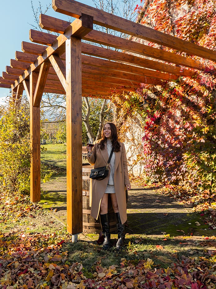 A woman in a tan coat and black boots enjoys a glass of wine while standing under a wooden pergola adorned with autumn foliage in Etyek, Hungary. Vibrant red and orange leaves cascade over a stone wall behind her, capturing the essence of the fall season. She smiles, embodying the relaxed atmosphere of a wine-tasting experience in a scenic vineyard setting.