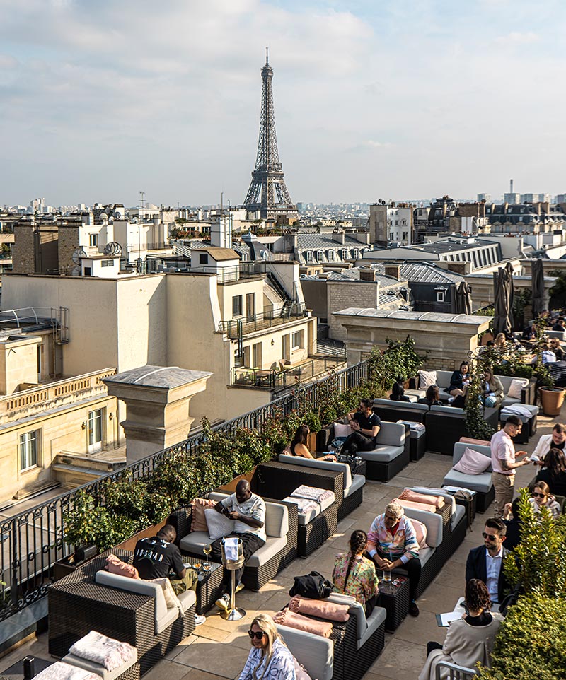 rooftop terrace filled with tables of people drinking cocktails with a view of the eiffel tower in the background