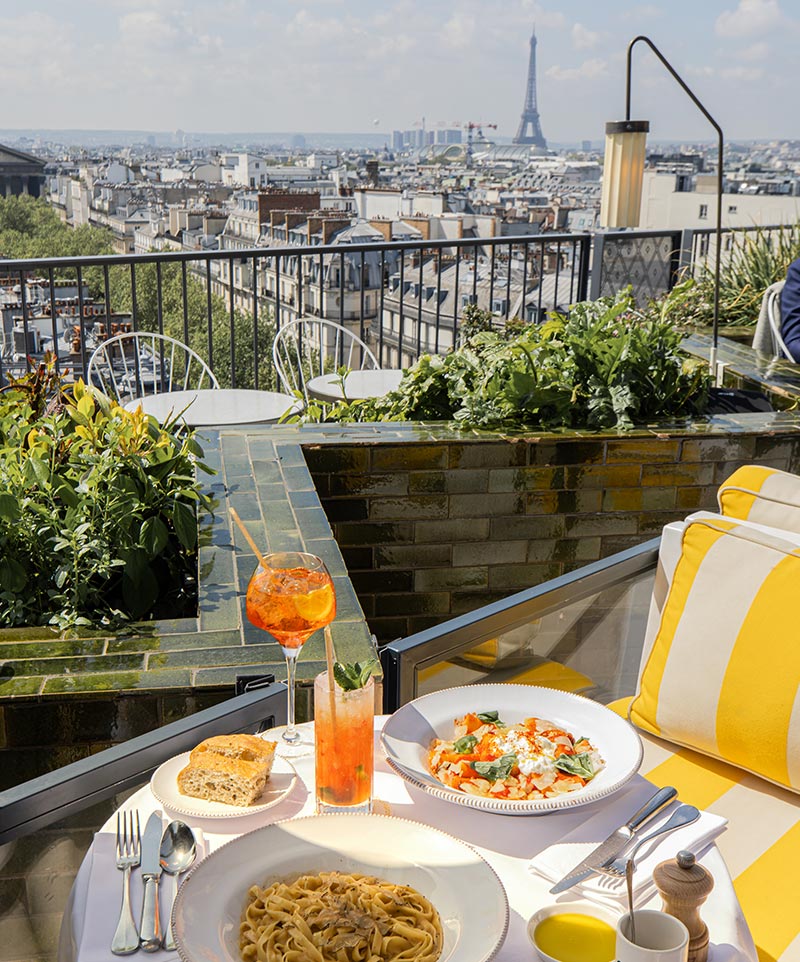 A rooftop dining table at Perruche in Paris set with a delicious meal, including a plate of pasta, a dish of gnocchi, a piece of focaccia bread, and two refreshing cocktails. The table is decorated with bright yellow and white cushions, enhancing the vibrant atmosphere. In the background, there is a stunning view of the Parisian skyline, with the Eiffel Tower prominently visible, creating a picturesque and memorable dining experience.


