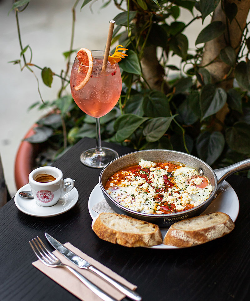 grapefruit cocktail on table with a cup of espresso next to a pan of shakshuka (baked eggs)