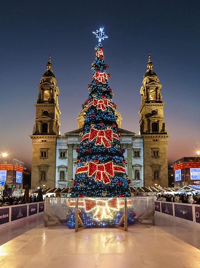 Alt text: A towering Christmas tree decorated with large red bows and blue lights stands illuminated in front of St. Stephen's Basilica in Budapest. The tree is topped with a glowing star, with an ice rink and bustling Christmas market booths surrounding it. The scene captures the festive atmosphere at twilight, with the basilica's twin towers providing a majestic backdrop.






