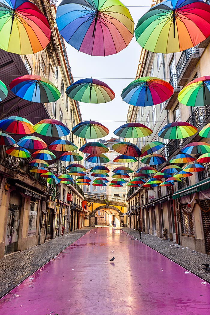 a street with a pink coloured ground, lined with bars and a ceiling of rainbow umbrellas