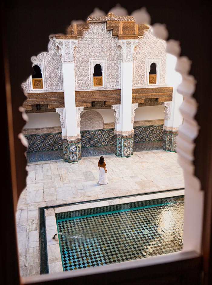 The image shows a view through an intricately carved archway, framing the inner courtyard of the Ben Youssef Madrasa in Marrakech, Morocco. A woman in a white dress stands near a patterned, turquoise-tiled pool, with the historic building's detailed mosaic walls and arches in the background. The scene captures the blend of traditional Moroccan architecture and tranquil ambiance.






