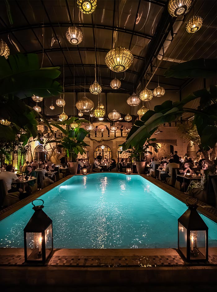 The image depicts an elegant dinner setting at La Trattoria in Marrakech, with a glowing turquoise pool at the center, surrounded by dining tables. The atmosphere is warm and inviting, enhanced by numerous hanging lanterns casting soft light across the space. Lush green plants and a blend of Moroccan and Mediterranean decor create a cozy, romantic ambiance, ideal for an intimate evening meal.


