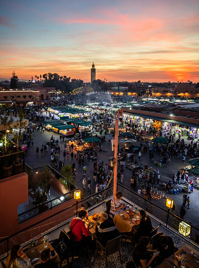 The image captures the lively evening atmosphere of Jemaa el-Fnaa square in Marrakech, viewed from a rooftop terrace. People dine at the terrace overlooking the bustling market below, filled with food stalls, vendors, and visitors. The sunset paints the sky with shades of orange and purple, while the Koutoubia Mosque stands tall in the background, adding to the vibrant, dynamic scene of this famous cultural hub.






