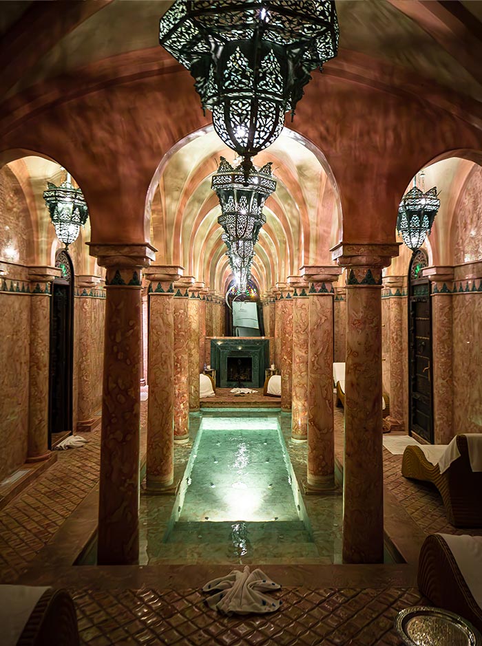 The image showcases the opulent interior of the spa at La Sultana in Marrakech, featuring a serene pool surrounded by warm-toned marble columns and arches. Elegant Moroccan lanterns hang from the ceiling, casting intricate shadows across the space, creating a tranquil and luxurious ambiance. The reflection of the water adds to the soothing atmosphere, making it an inviting setting for relaxation and rejuvenation.






