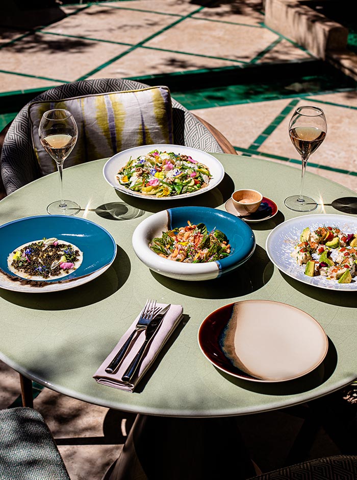 The image features an elegant outdoor dining setup at La Table, located at Royal Mansour in Marrakech. The round table is adorned with colorful dishes, including vibrant salads and artfully plated appetizers, accompanied by two glasses of wine. The scene is bathed in sunlight, highlighting the fresh ingredients and sophisticated ambiance of the space, with patterned cushions and a serene garden backdrop adding to the inviting atmosphere.