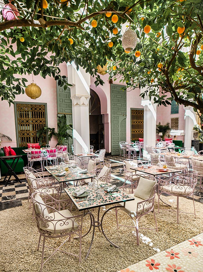 The image showcases the charming outdoor seating area of Les Jardins Du Lotus in Marrakech. The space is adorned with intricately designed tables and wrought iron chairs, cushioned for comfort, set beneath a leafy canopy of citrus trees laden with bright yellow lemons. The courtyard’s pastel pink walls and green doors add to the serene, inviting atmosphere, making it a perfect spot for a relaxed meal in a lush, garden-like setting.