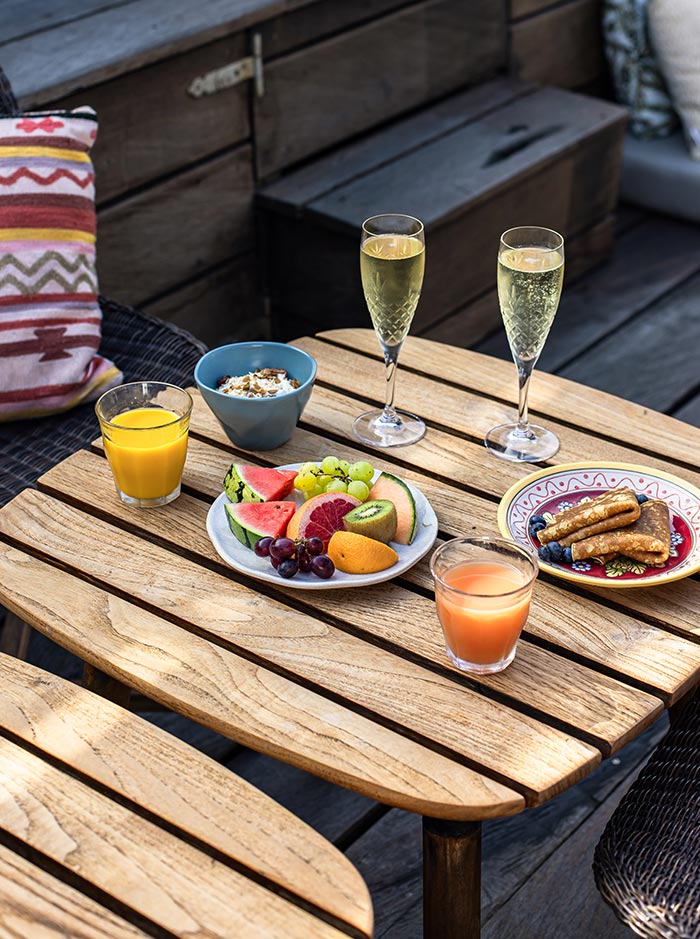 The image shows a delightful breakfast setup at Manon Les Suites in Copenhagen. A wooden table is adorned with plates of fresh fruit, a bowl of yogurt topped with granola, a dish of pancakes, and glasses of orange and grapefruit juice. Two flutes of sparkling wine add a touch of indulgence to the meal. The cozy setting is complemented by a patterned cushion and rustic wooden benches, creating a warm and inviting atmosphere perfect for a leisurely morning.