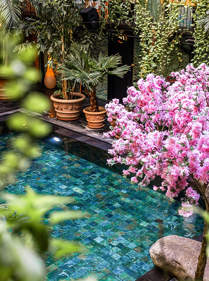 The image depicts a lush indoor pool area at Manon Les Suites in Copenhagen, surrounded by abundant greenery. The pool's turquoise tiles shimmer under the light, creating a serene atmosphere. Vibrant pink blossoms from a nearby tree add a pop of color, while potted palm plants and cascading vines contribute to the tropical, oasis-like setting. The combination of natural elements and tranquil water creates a peaceful retreat in the heart of the hotel.