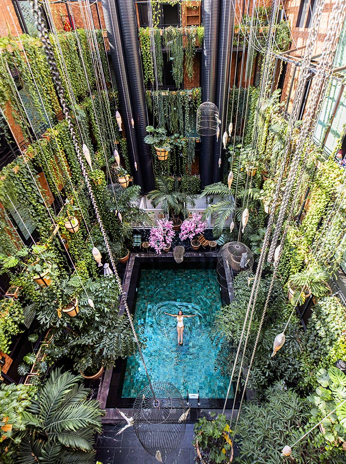 The image captures the stunning indoor pool area at Manon Les Suites in Copenhagen. The pool is surrounded by lush greenery, with plants cascading down from multiple floors, creating a vertical garden effect. Hanging lanterns and birdcage-inspired decorations add an eclectic, bohemian touch to the space. A swimmer enjoys the serene atmosphere in the turquoise water, framed by vibrant foliage and exotic plants, making it feel like a hidden oasis in the heart of the city.