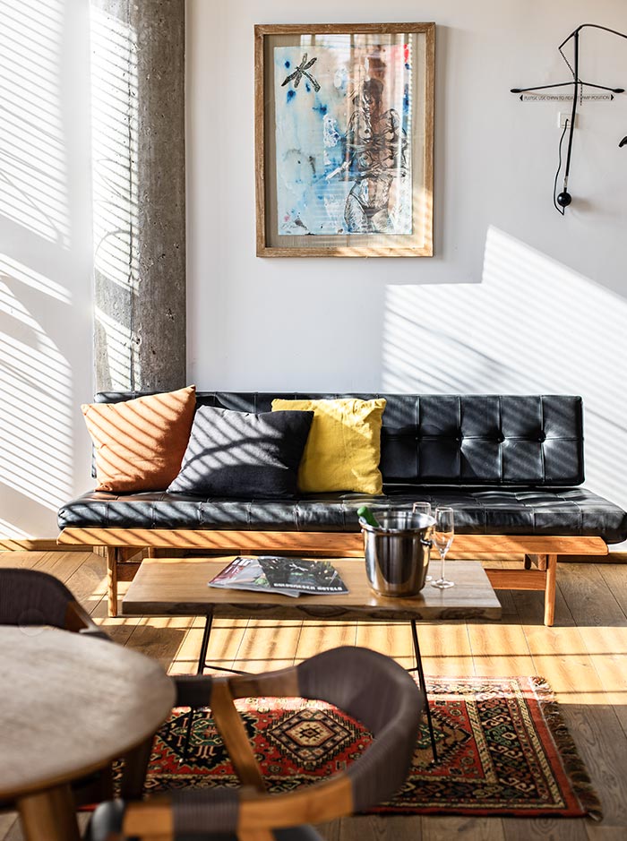 The image captures a cozy, sunlit corner of a room at Manon Les Suites in Copenhagen. A black tufted sofa adorned with colorful cushions—yellow, orange, and dark gray—sits against a white wall. Above it hangs a framed artwork, adding a touch of character to the space. A wooden coffee table holds a chilled bottle of champagne in an ice bucket with two flutes, inviting relaxation. The warm sunlight filters through blinds, casting soft shadows on the floor and highlighting the vibrant patterned rug that adds a cozy and eclectic vibe to the room.