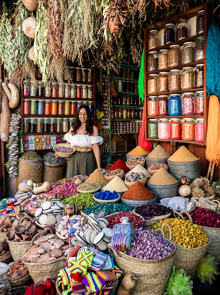 The image shows a vibrant scene at a spice and herb stall in Souk Semmarine, Marrakech. A woman in a white off-the-shoulder top stands amidst baskets filled with colorful spices, dried flowers, and natural dyes, holding a bowl of dried lavender. Behind her, shelves are lined with jars of herbs, and bundles of dried plants hang from above, creating a rich, sensory display of Moroccan market life.






