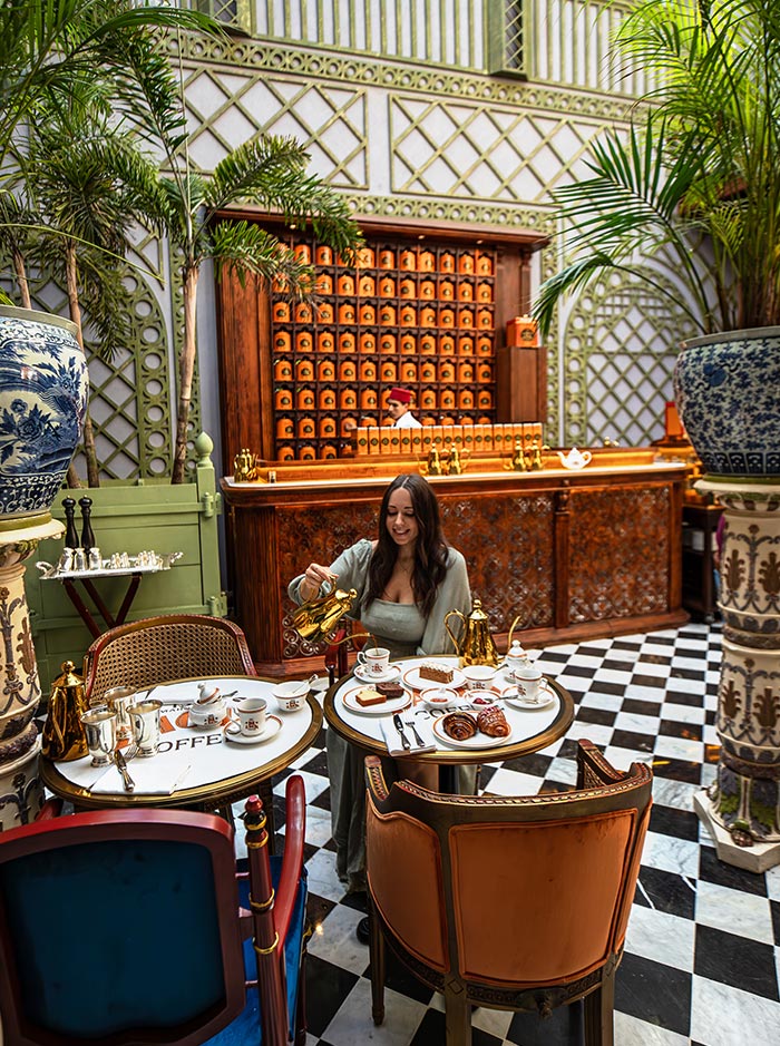 The image shows a woman pouring coffee from a golden pot at Bacha Coffee in Marrakech. She is seated at a small table adorned with cups, saucers, pastries, and a variety of coffee accouterments, set against a luxurious backdrop with a checkerboard floor, ornate wooden counter, and lush potted plants. The scene exudes a blend of elegance and relaxation, capturing the sophisticated ambiance of the café.


