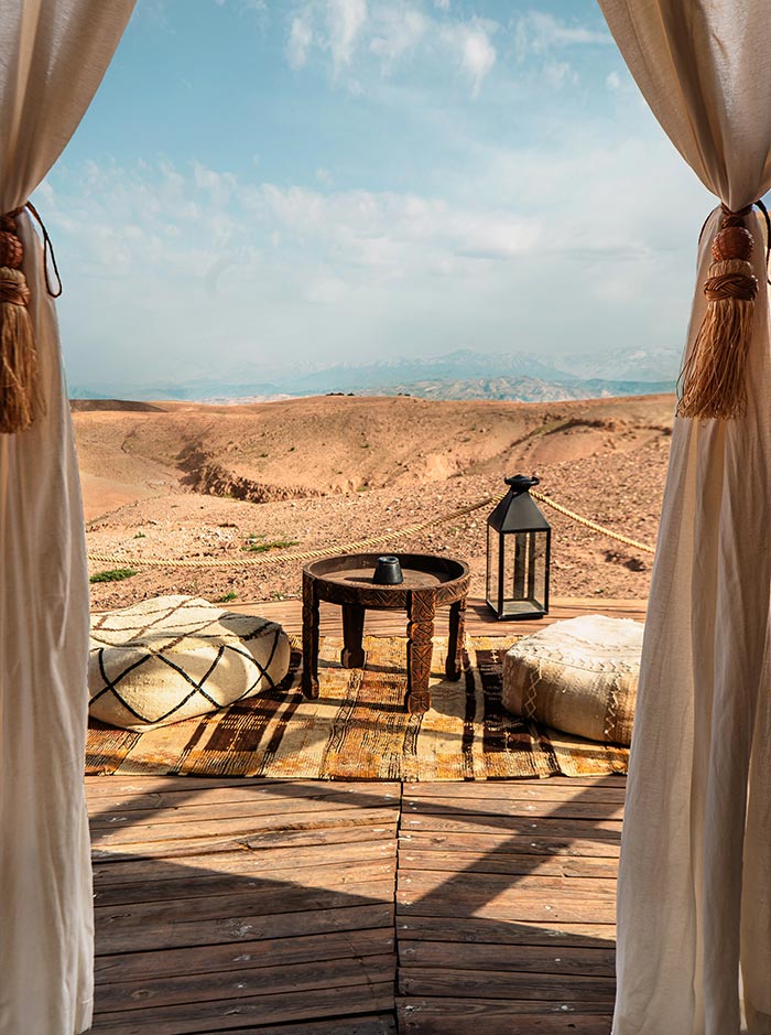 The image captures a peaceful scene from inside a tent at a desert camp in the Agafay Desert near Marrakech. The view looks out over arid, rolling hills and distant mountains, framed by cream-colored curtains. In the foreground, a small wooden table, a lantern, and cozy poufs rest on a woven rug, creating an inviting space to relax and enjoy the vast desert landscape.






