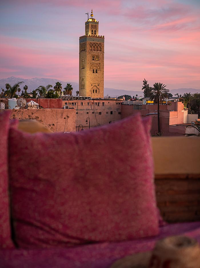 The image features a stunning view of the Koutoubia Mosque at sunset from the rooftop of El Fenn in Marrakech. The sky is painted with shades of pink and purple, providing a beautiful backdrop to the illuminated mosque and the surrounding cityscape. In the foreground, a cozy seating area with pink cushions adds warmth and comfort, framing the iconic landmark against the serene evening sky.