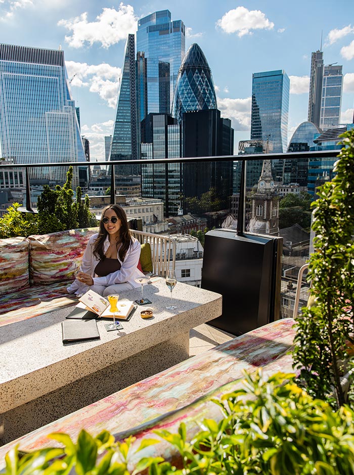 A woman enjoys a drink at Florattica Rooftop Bar in London, seated on a colorful bench with a table holding menus and drinks. The skyline in the background includes the distinctive Gherkin building and other skyscrapers, offering a stunning view of the city's modern architecture under a clear blue sky.