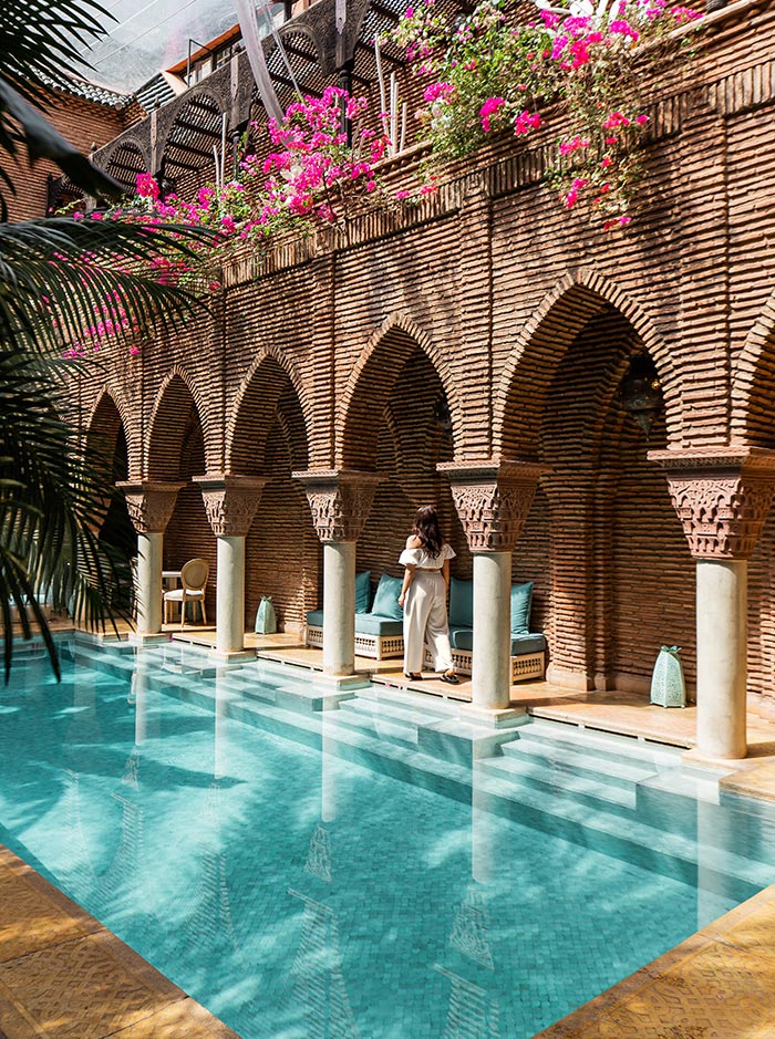 The image shows a luxurious poolside scene at La Sultana in Marrakech, featuring a turquoise pool surrounded by arched brick walls and columns. A woman stands near the edge, looking out towards the pool, with comfortable seating areas tucked into the arches behind her. Bright pink bougainvillea flowers cascade from above, adding a vibrant touch to the serene and elegant ambiance of this Moroccan oasis.