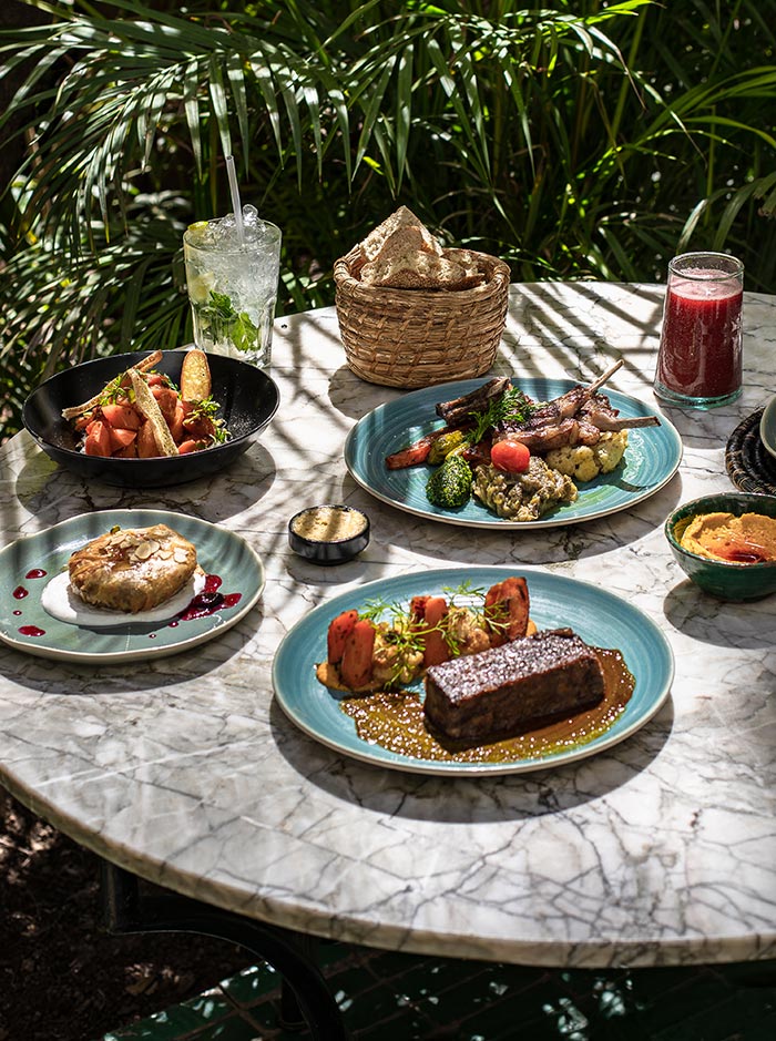 The image displays a beautifully arranged table at Le Jardin restaurant in Marrakech, set outdoors amidst lush greenery. The marble table is topped with a variety of dishes, including grilled meats, roasted vegetables, bread in a wicker basket, and vibrant drinks like a mojito and a berry smoothie. The scene highlights the freshness and variety of Moroccan cuisine in a serene, natural setting.






