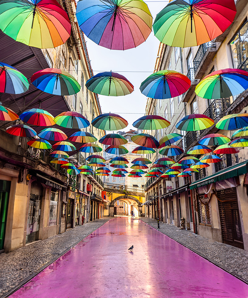 a pink coloured street with rainbow coloured umbrellas suspended above 