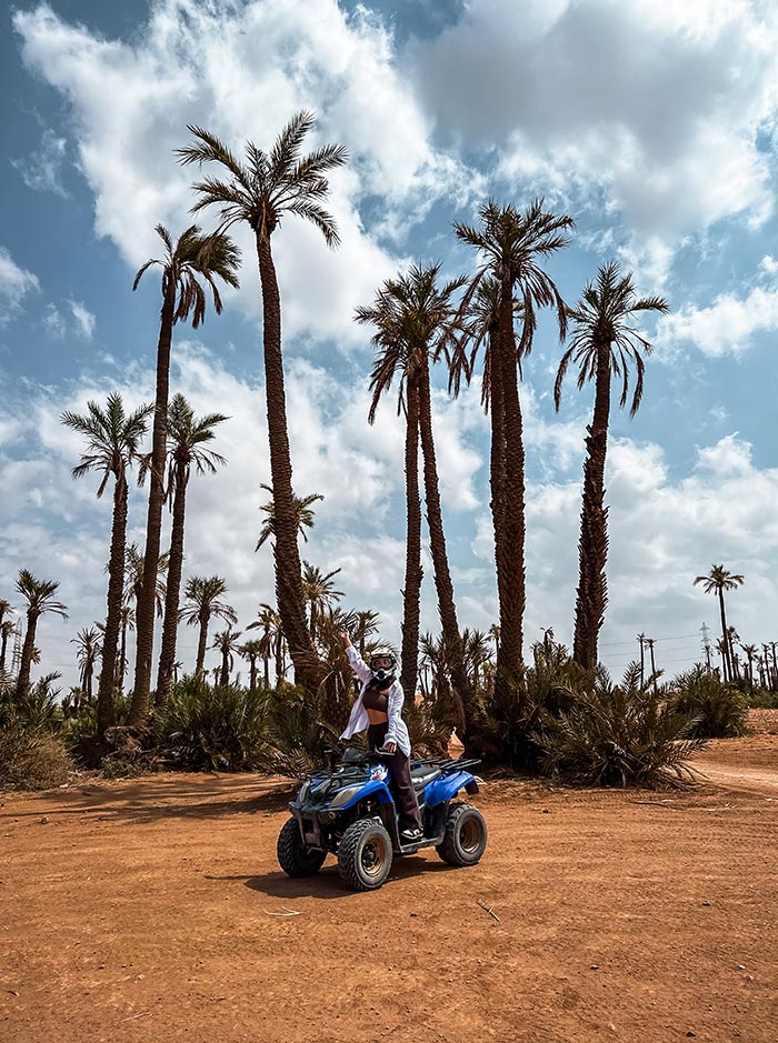 The image shows a person on a blue ATV, wearing a helmet and goggles, posing with one arm raised amidst the palm trees of the Marrakech desert. The scene captures the adventurous spirit of quad biking, set against a backdrop of tall palms and a bright, partly cloudy sky. The sandy terrain and rugged landscape highlight the thrill of exploring the outdoors in this desert environment.







