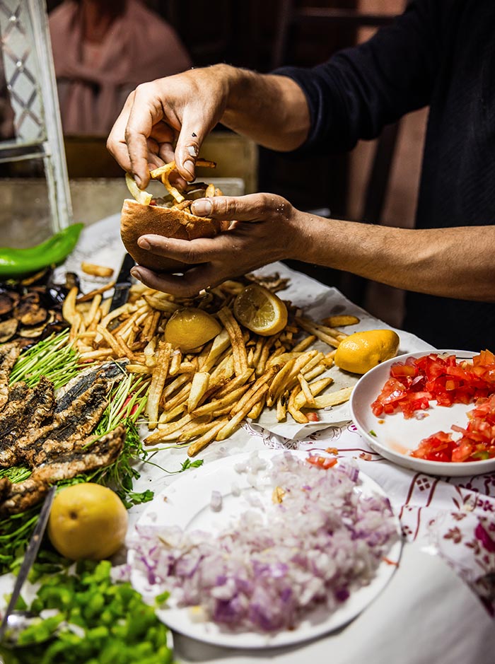 The image captures a scene from a Marrakech street food stall, where a vendor is preparing a sandwich. The table is filled with fresh ingredients like fried fish, golden French fries, lemon halves, chopped tomatoes, and onions. The vendor’s hands are in action, assembling the sandwich amidst a vibrant spread, highlighting the richness and diversity of Moroccan street food.






