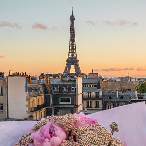 A picturesque view of the Eiffel Tower at sunset, framed by charming Parisian rooftops. The sky is painted in soft pastel hues of orange and pink, adding a romantic ambiance. In the foreground, a bouquet of pink flowers wrapped in white paper adds a delicate touch, enhancing the overall elegance of the scene. The iconic Eiffel Tower stands tall and majestic in the background, creating a quintessentially Parisian moment.






