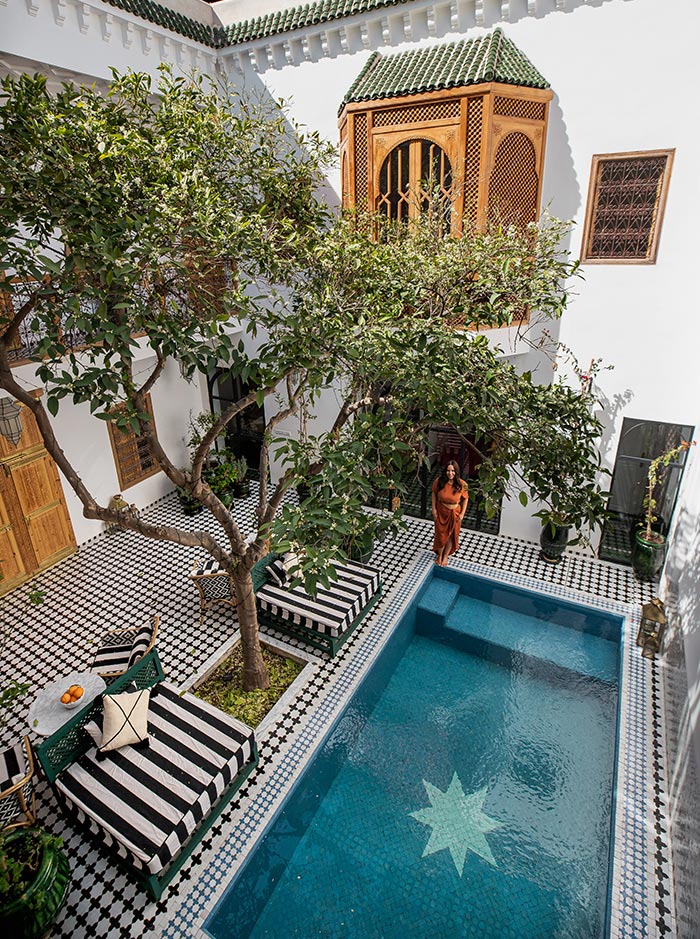 The image captures the serene courtyard of Riad Botanica in Marrakech, featuring a turquoise pool with a star design at the bottom. The space is adorned with striped lounge chairs, a small round table, and lush greenery, including trees that provide shade. A woman in a rust-colored dress stands near the pool, adding a touch of warmth to the elegant setting, highlighted by white walls and green-tiled roof details.






