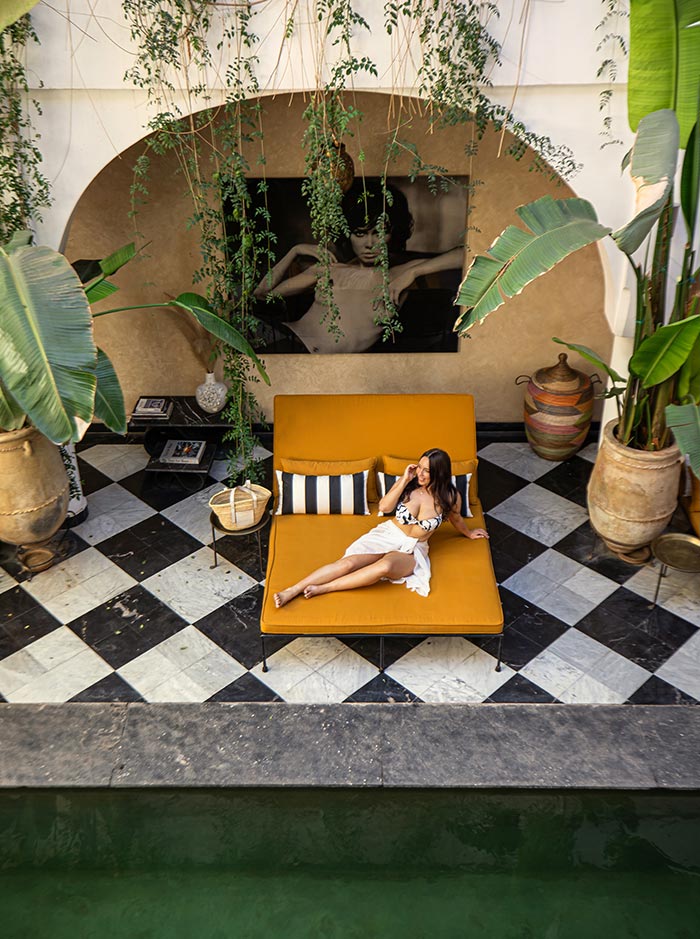 The image showcases a stylish courtyard at Riad Sakkan, featuring a woman lounging on a mustard-yellow daybed beside a small pool. The space is decorated with lush hanging plants and large potted greenery, creating a serene atmosphere. Black-and-white checkered tiles and a framed black-and-white photograph on the wall add a chic, modern touch to the traditional Moroccan ambiance.