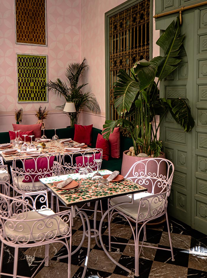 The image features a beautifully set dining area at Les Jardins Du Lotus in Marrakech. The space includes a mosaic-topped table surrounded by pastel pink wrought iron chairs with cushioned seats. The decor is elegant, with vibrant pink and green accents, including plush pillows, large potted plants, and artistic wall panels, all under soft lighting that complements the warm, inviting atmosphere.