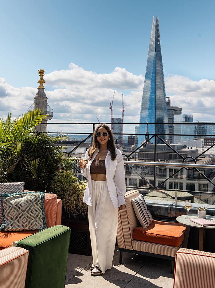 A woman stands on the terrace of Wagtail Rooftop Bar in London, holding a drink and smiling. The background features a stunning view of The Shard and other cityscape landmarks. The terrace is furnished with colorful, comfortable seating and lush plants, creating a chic and inviting atmosphere under a partly cloudy sky.