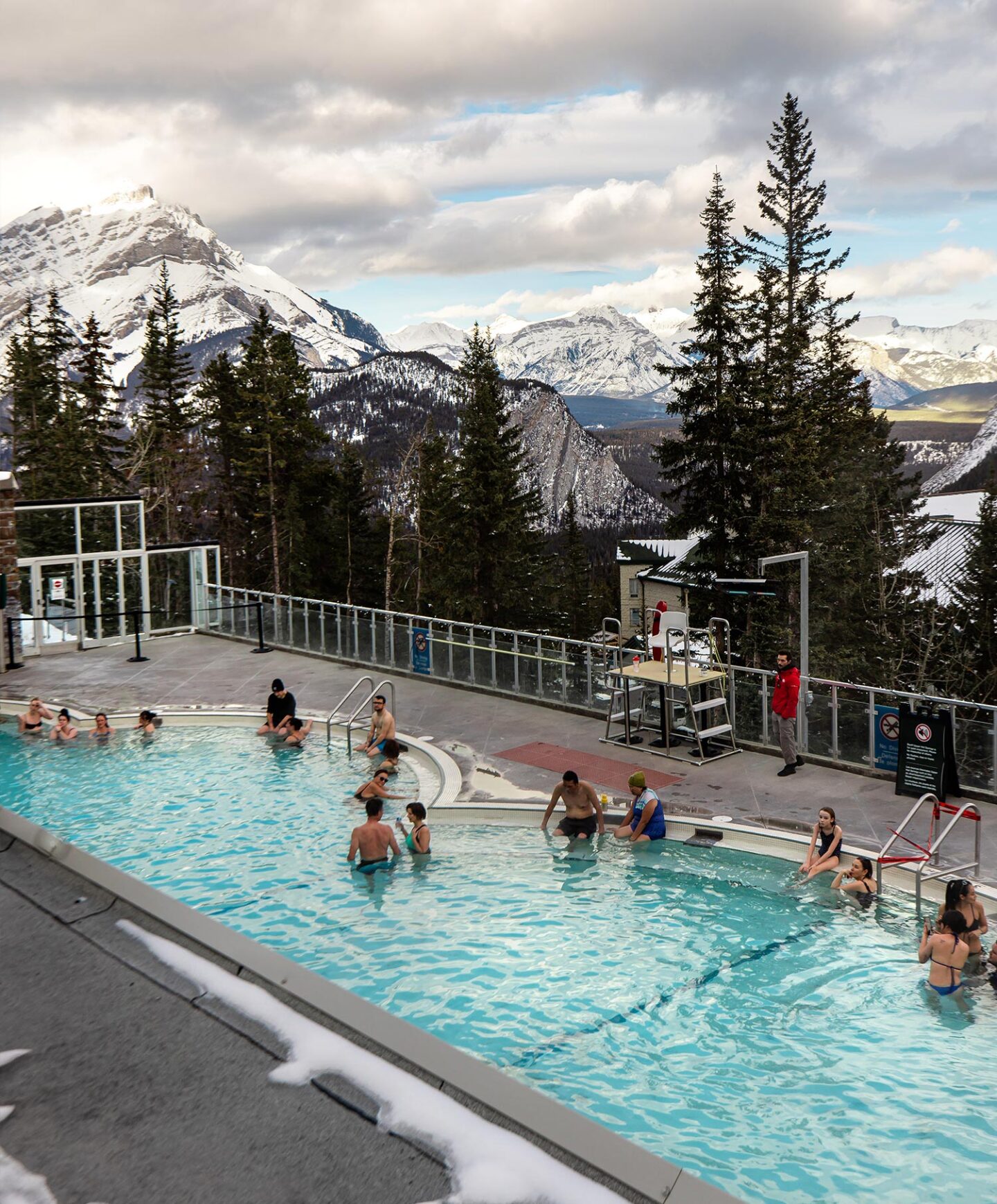 A small group of people are bathing in a natural hot spring pool, from the balcony of this pool you can see snow capped mountains in the background