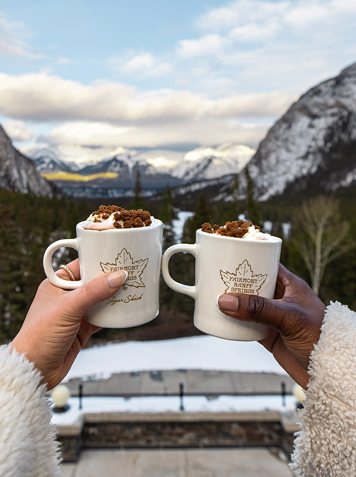 Two hands are holding mugs of hot chocolate, Behind the hands is a view of snow capped mountains 