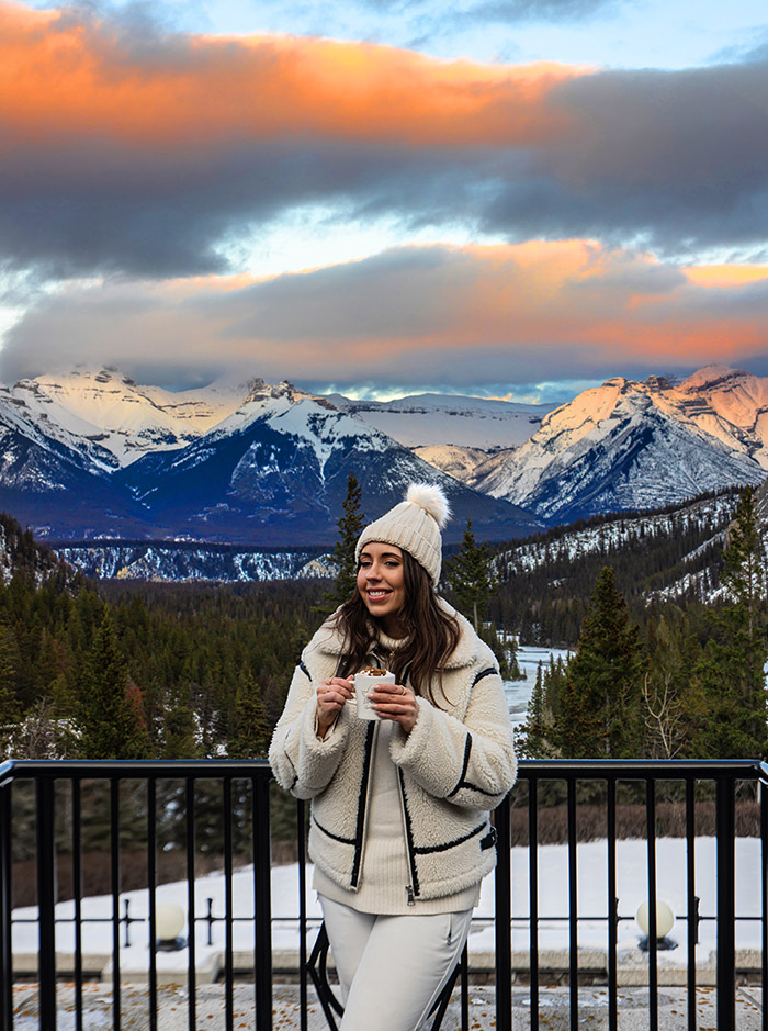 Kelsey is wearing a beanie and winter jacket, holding a mug of hot chocolate. Behind her is a view of snow capped mountains and a sunset sky