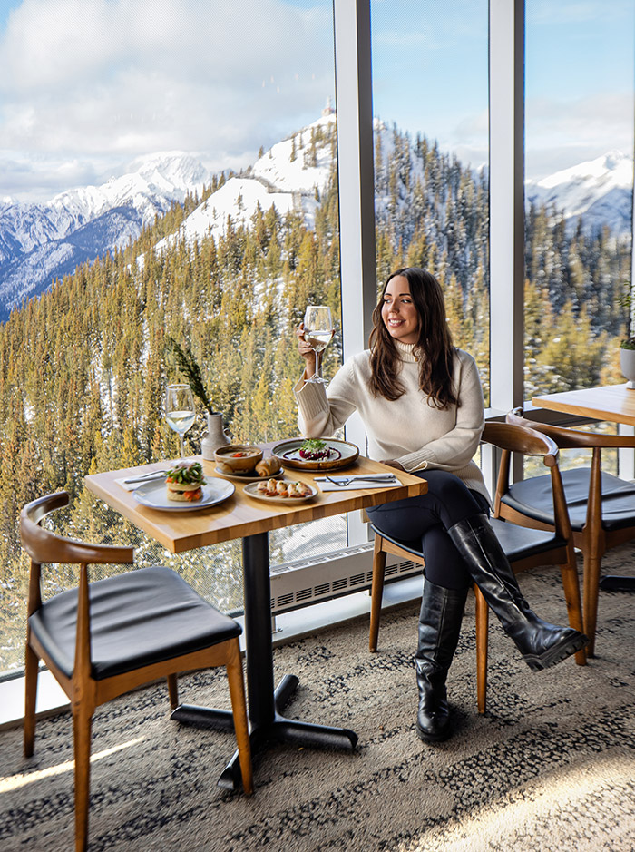 Kelsey sitting at a table for two, she is holding a glass of wine and enjoying views of snow capped mountains from the restaurant window