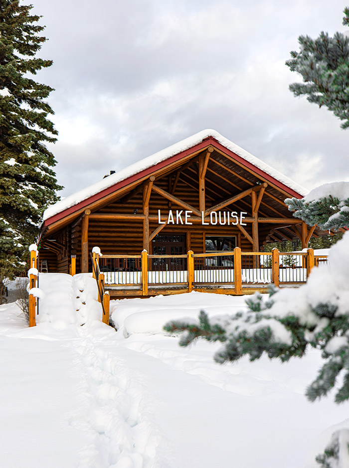 A wooden restaurant with the sign "Lake Louise", the restaurant is surrounded by heavy snowfall and pine trees